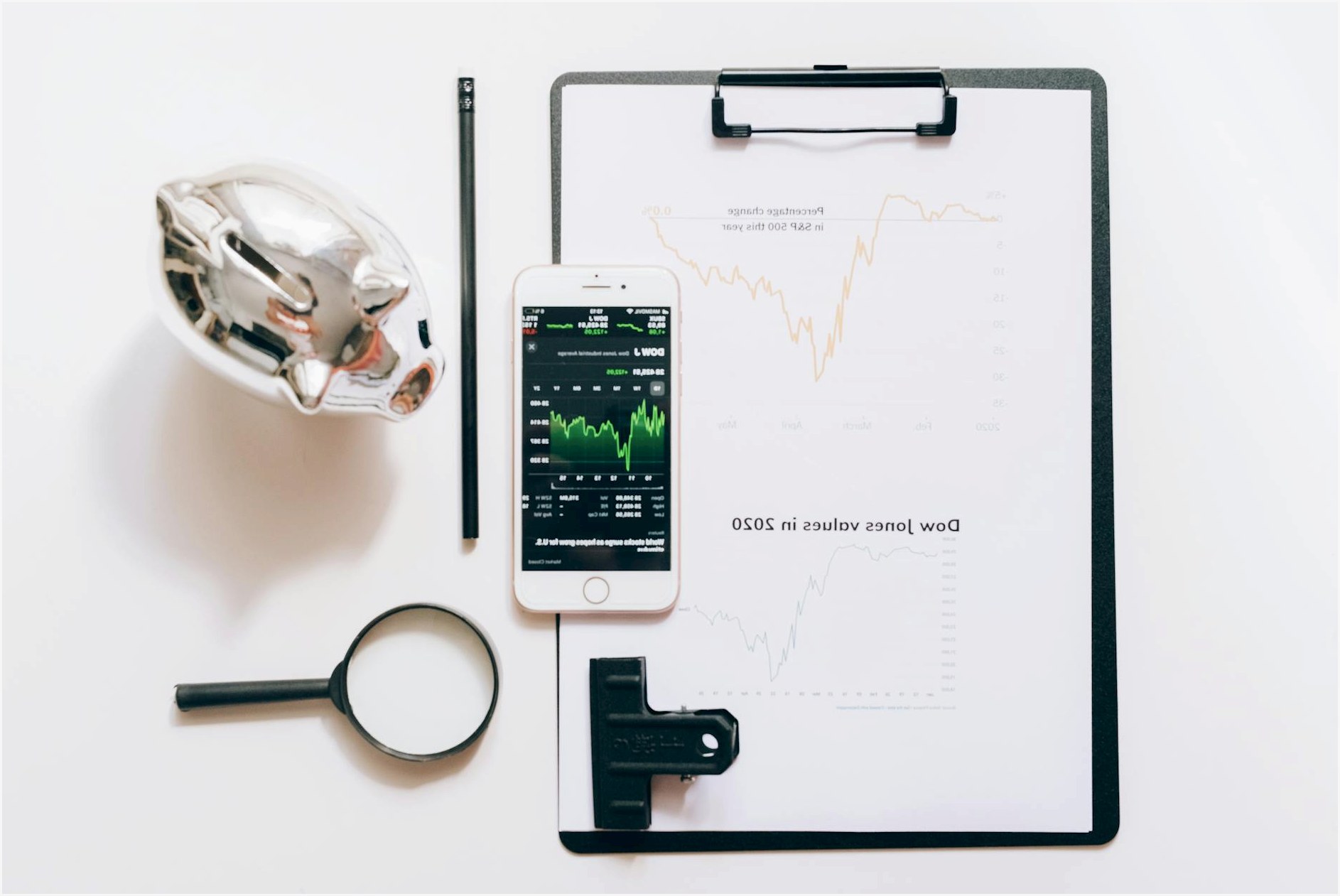 Top view of financial analysis tools including a cellphone, clipboard, magnifying glass, and piggy bank on a white desk.