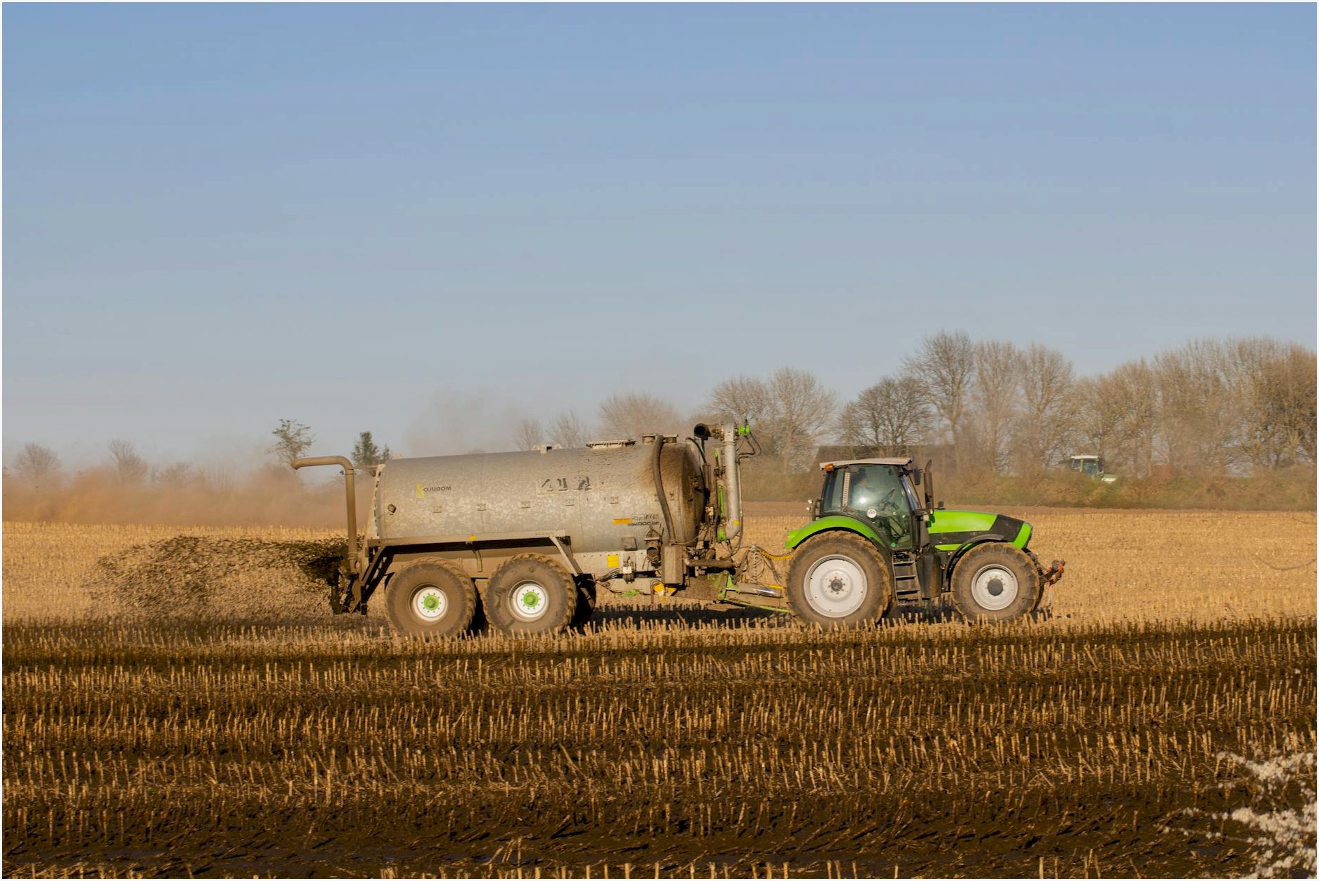 A tractor with a tanker spreading fertilizer over a barren farmland under clear skies.