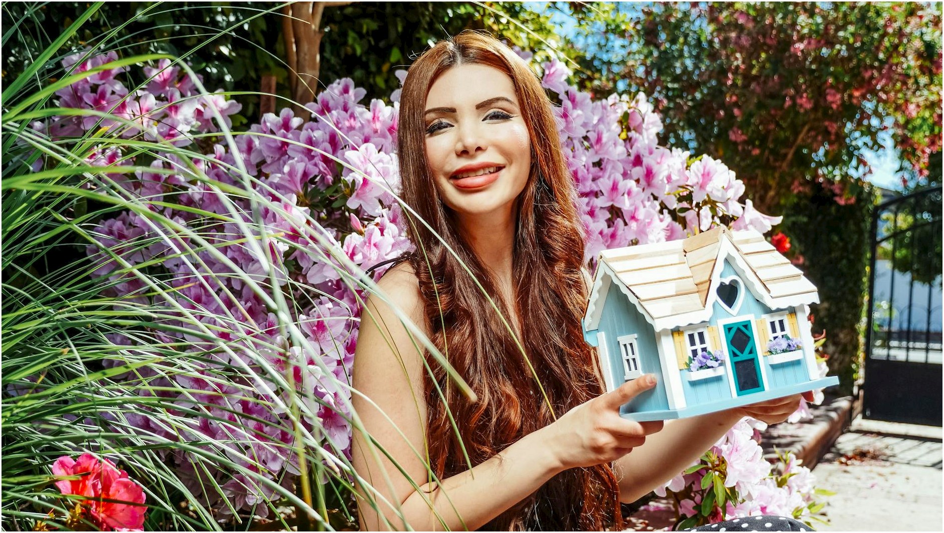 A woman holding a tiny house model among vibrant flowers in a sunny garden scene.