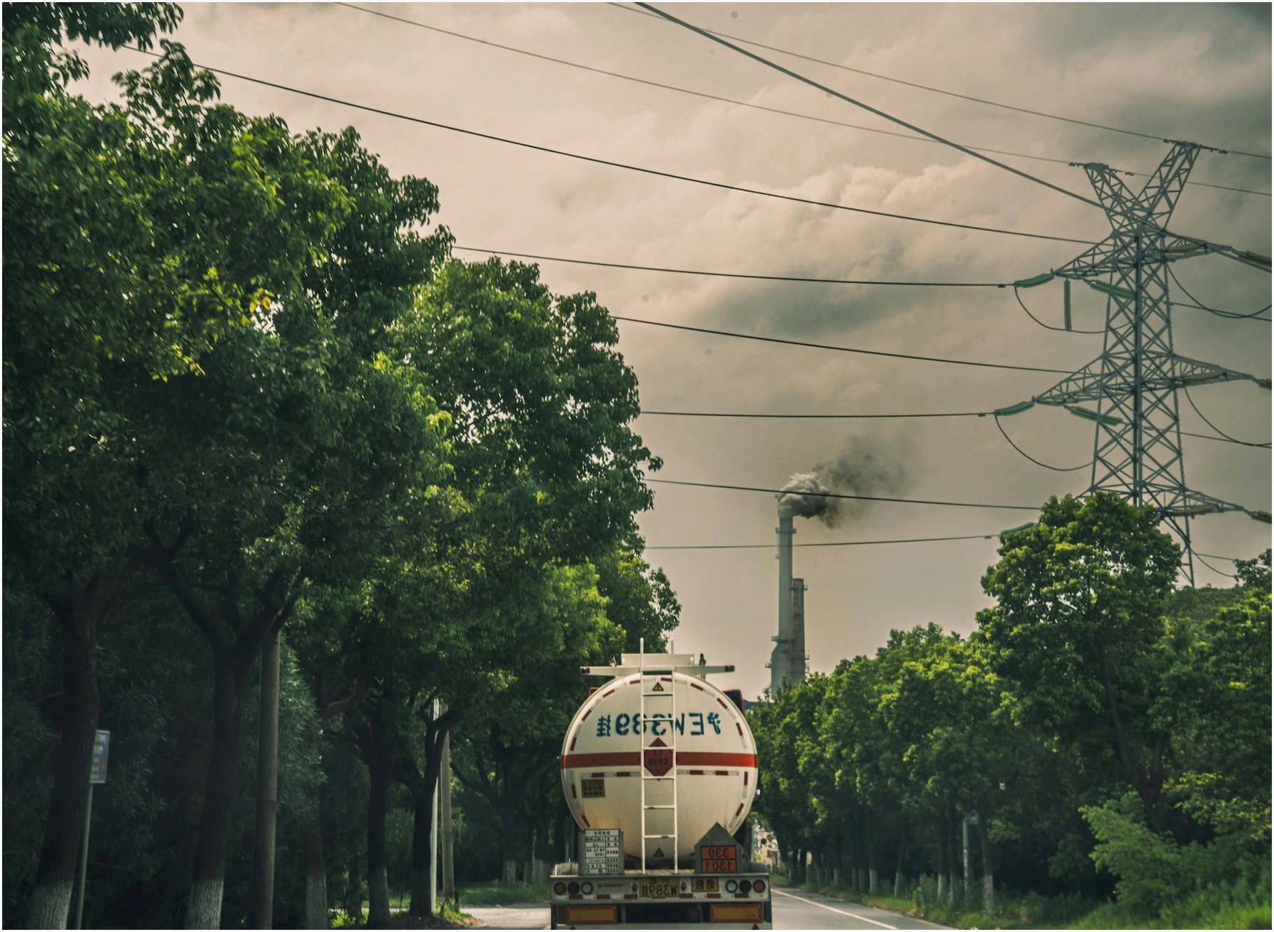 Tanker truck driving on a road by a smokestack emitting smoke under power lines and trees.