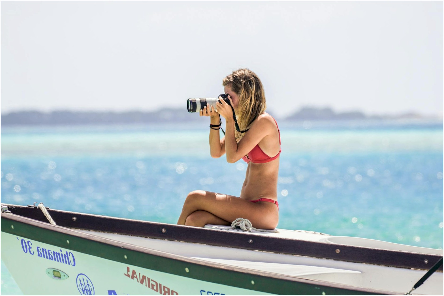 A woman in a bikini sits on a boat, taking photos of the scenic ocean view.