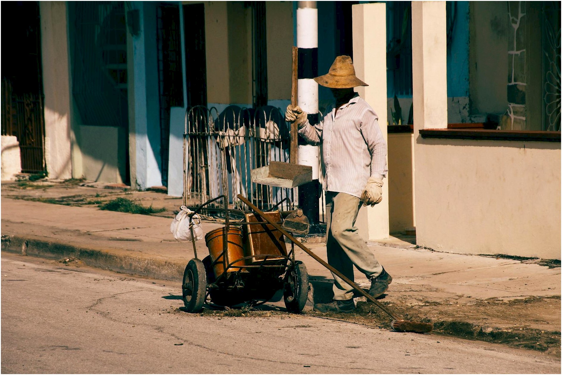 A street cleaner wearing a hat and shirt is working on a sidewalk with a cart in an urban setting.