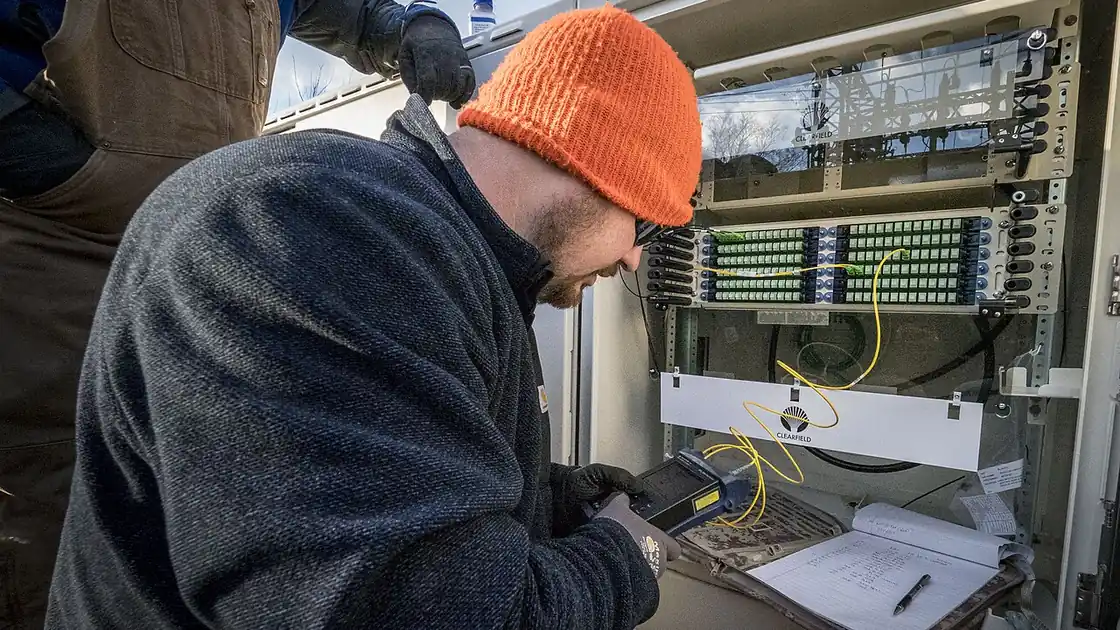 Técnico testa conexão de fibra óptica em equipamento de rede; imagem contextual sobre instalação de internet rural.
